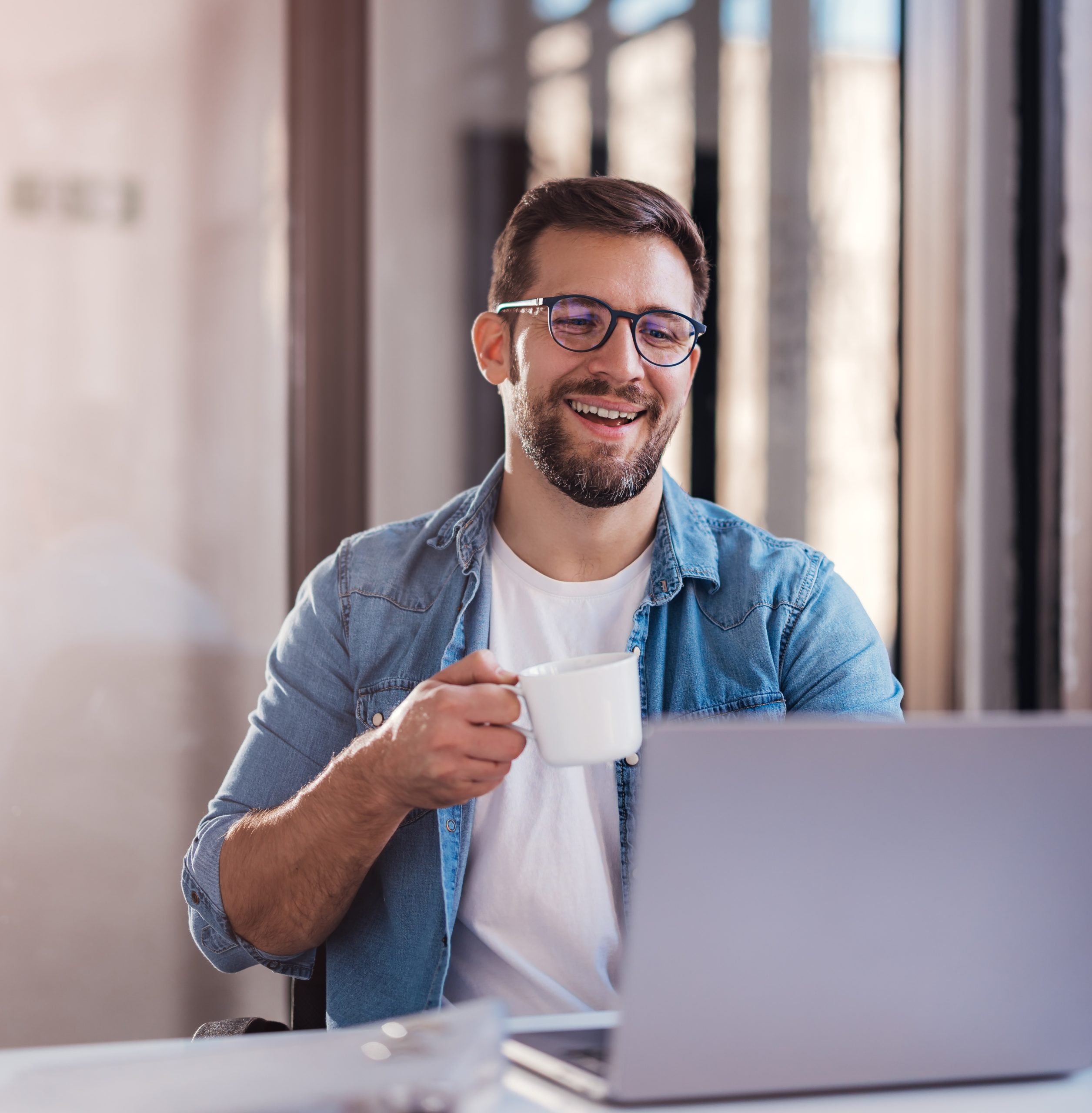 Handsome smiling young man sitting by window in office, drinking coffee while working on laptop.