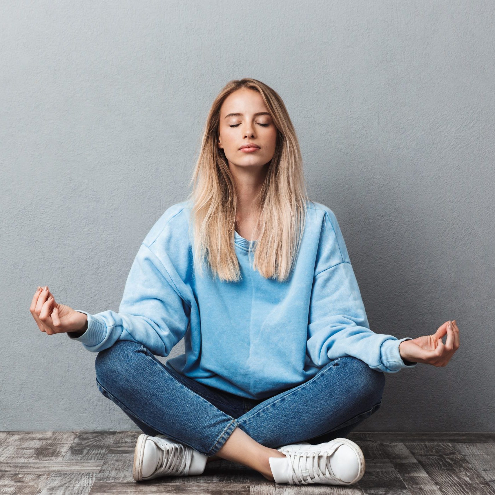 Pretty young blonde girl meditating while sitting in a yoga position with eyes closed isolated over gray background