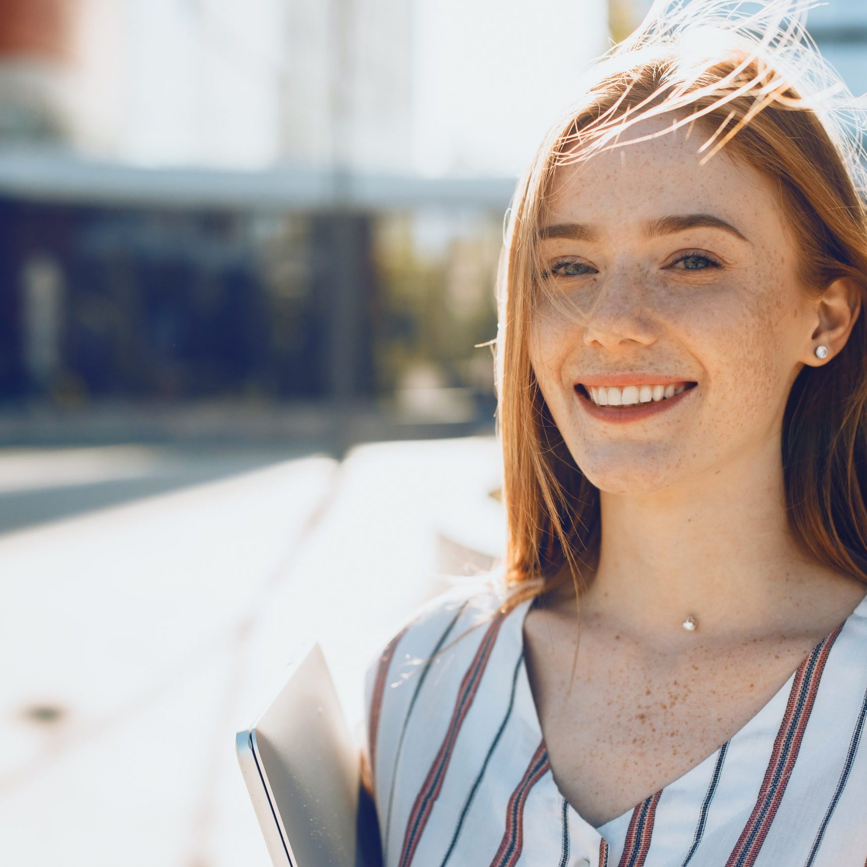 Close up portrait of a cute young woman with red hair and freckles looking at camera smiling against sunset holding a laptop .