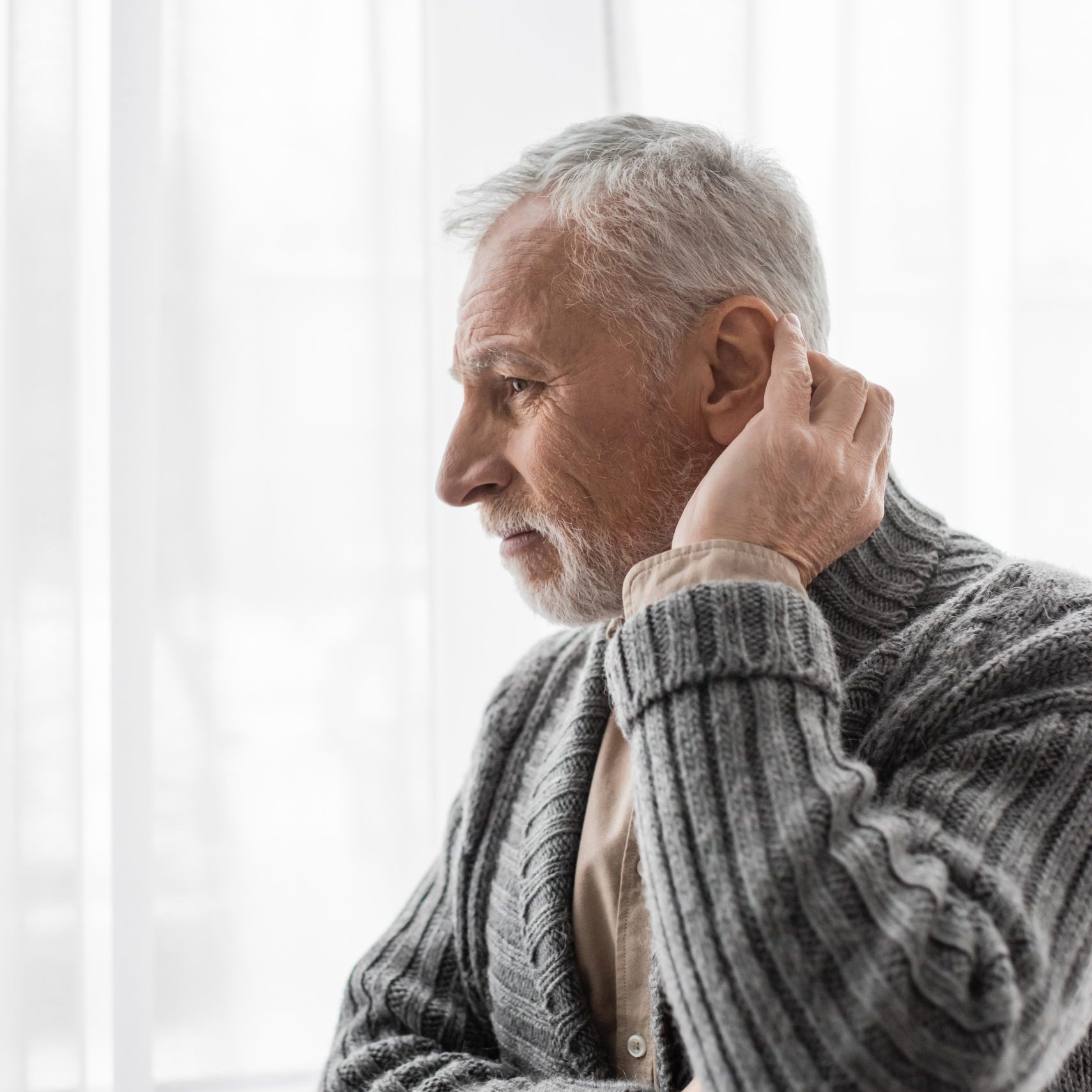 senior and thoughtful man in knitted cardigan looking away near window while suffering from alzheimer syndrome,stock image